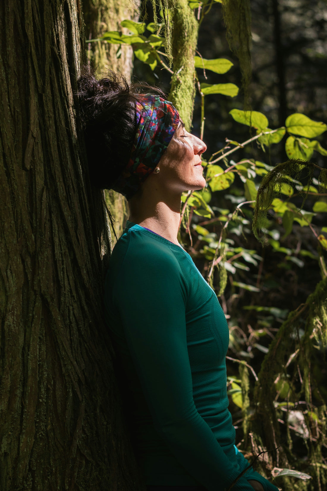 A woman sat with her eyes closed supported by a tree trunk and gazing towards the sun while sat in a leafy undergrowth. 